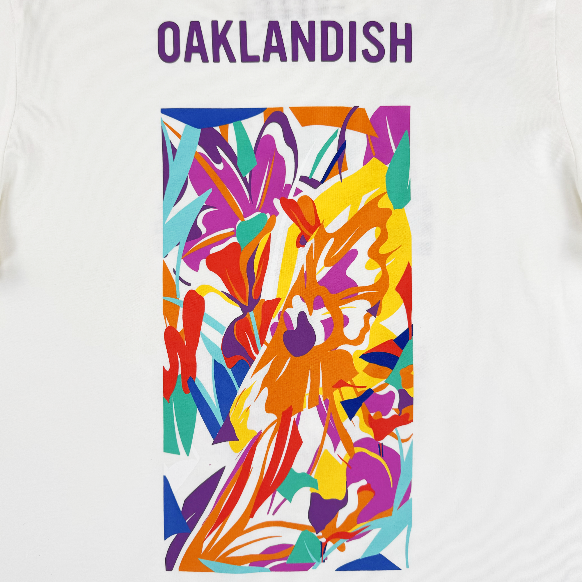 The Oaklandish Urban Tropics Tee features bold purple "OAKLANDISH" text above Kalani Ware’s vibrant abstract art with overlapping shapes in red, orange, yellow, teal, blue, and magenta for a dynamic look.