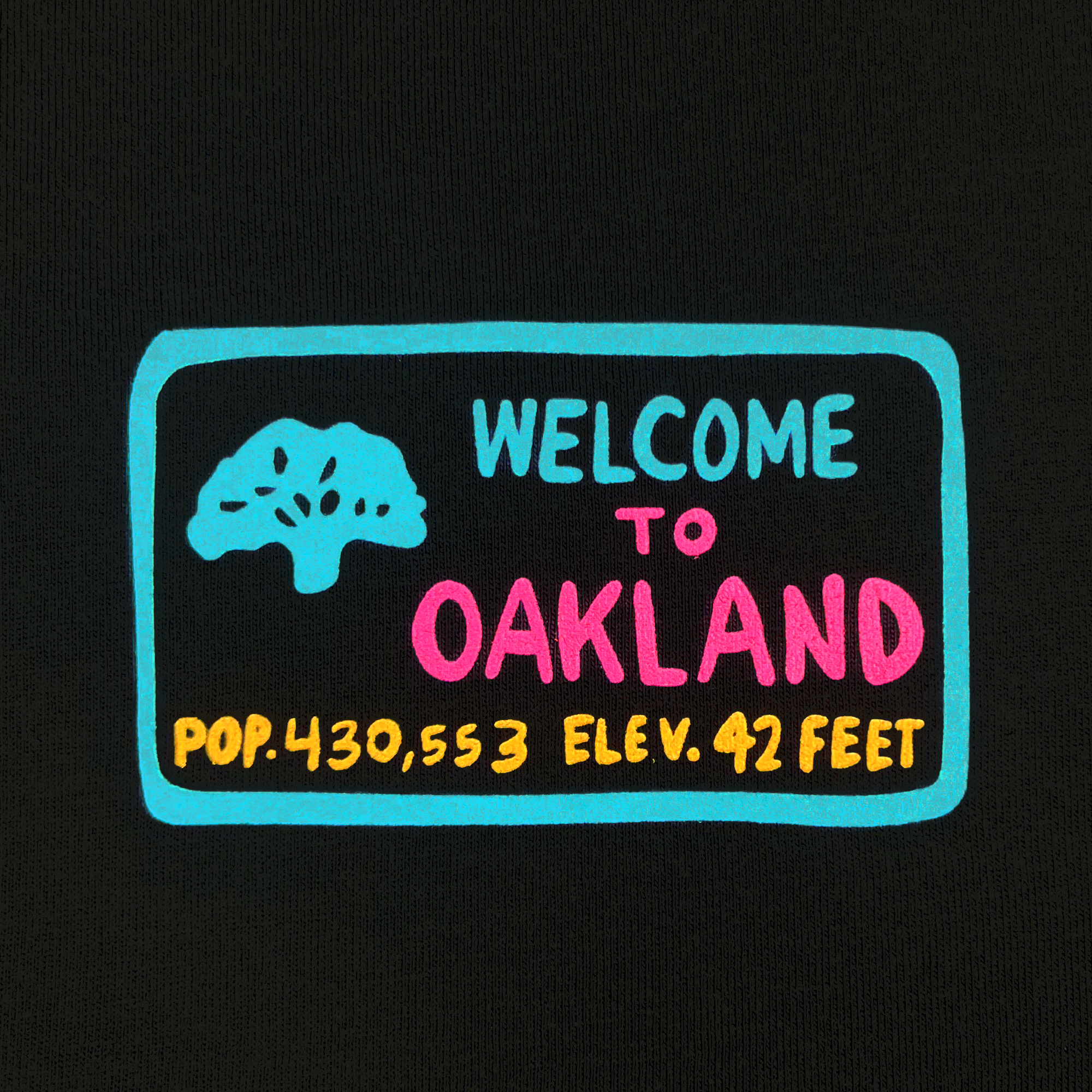 The Tune Town Tee from Oaklandish showcases a black backdrop adorned with a graphic featuring the text "WELCOME TO OAKLAND." Above the text is a light blue stylized tree icon, while below it states "POP. 430,553 ELEV. 42 FEET" in orange and yellow hues. The word "WELCOME" appears in cyan and "OAKLAND" in pink, encapsulating an authentic Oaklandish vibe.