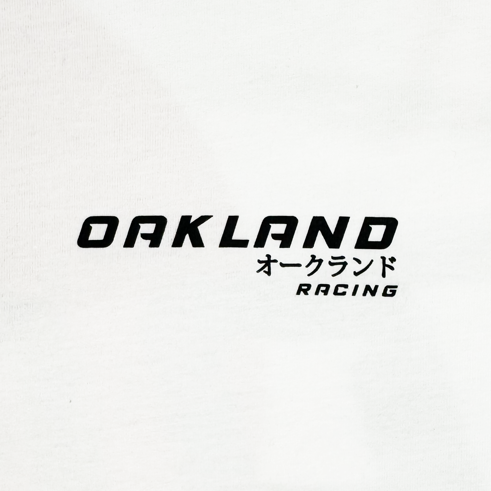 The Tuner LS Tee from Oaklandish is a white, 100% cotton long sleeve shirt with bold black "OAKLAND" in caps and Japanese katakana "オークランド." Below, "RACING" appears in smaller letters, slightly tilted right. It offers a classic fit with a modern and simple design.