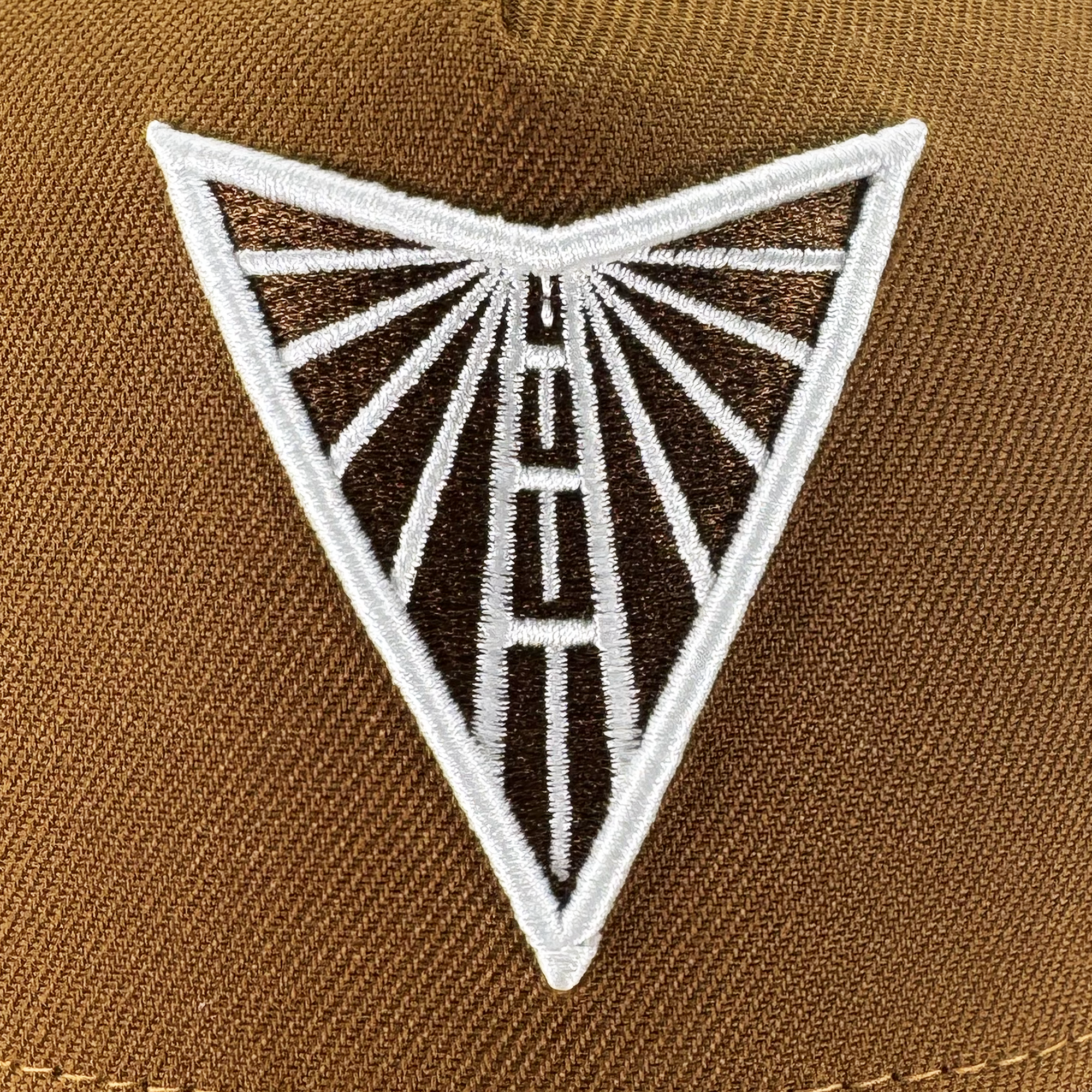 A close-up of the GSV Logo 9FORTY AF Snapback by GSV shows a brown adjustable cap with a triangular patch featuring a white and black design of lines radiating from a central tower.