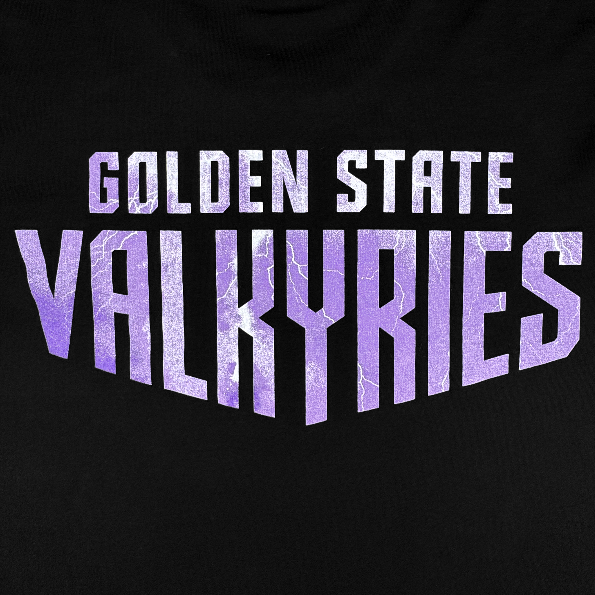 The GSV Golden State Valkyries Violet Lightning LS Tee features bold white "GOLDEN STATE" text above large purple "VALKYRIES" letters with a lightning texture on organic cotton, capturing the energy of the team in a striking design.