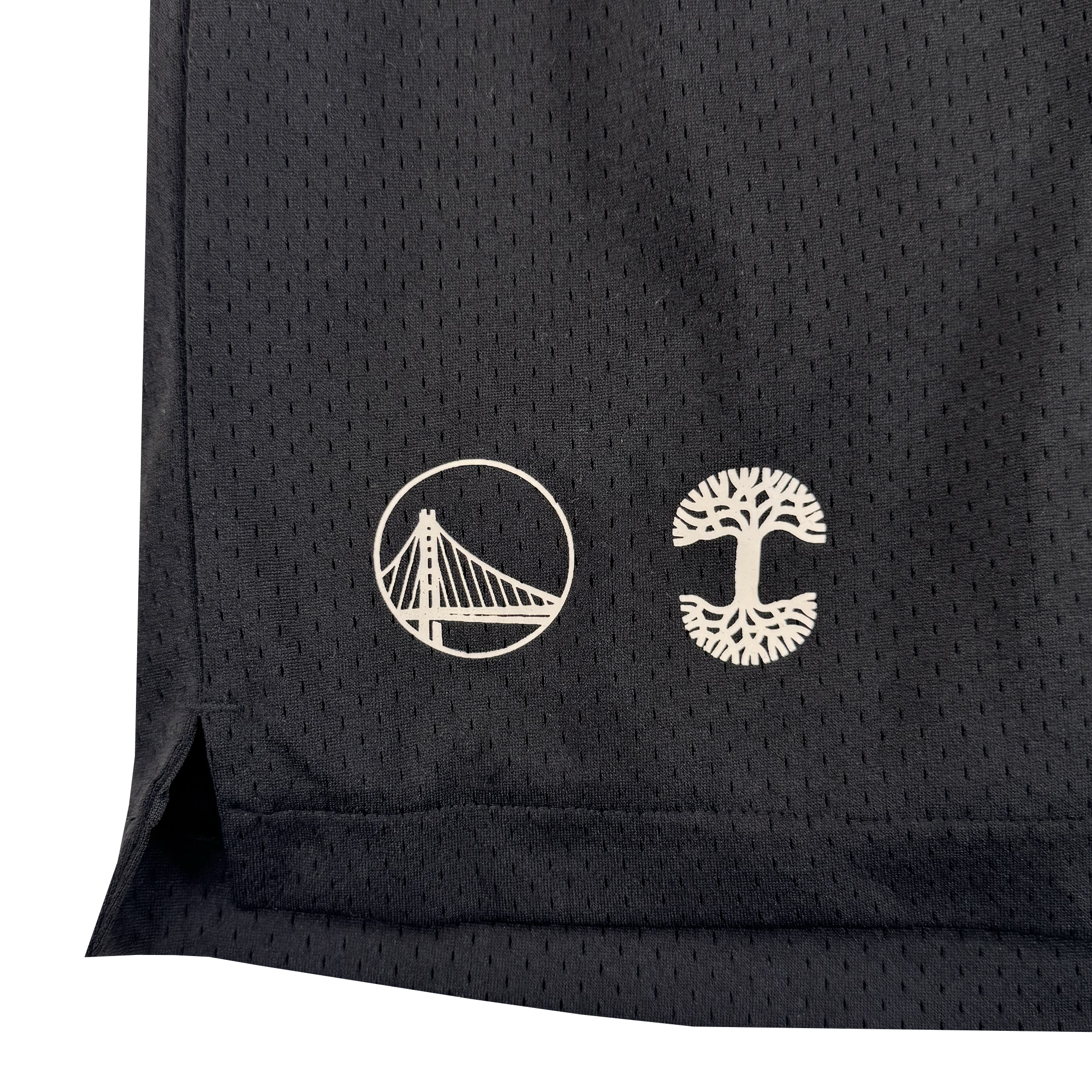 A close-up of black athletic mesh fabric on the GSW Arched Black-Letter Shorts shows two white logos—a suspension bridge in a circle and an Oaklandish tree. As part of the Town Collection, the shorts feature a small side slit at the edge.