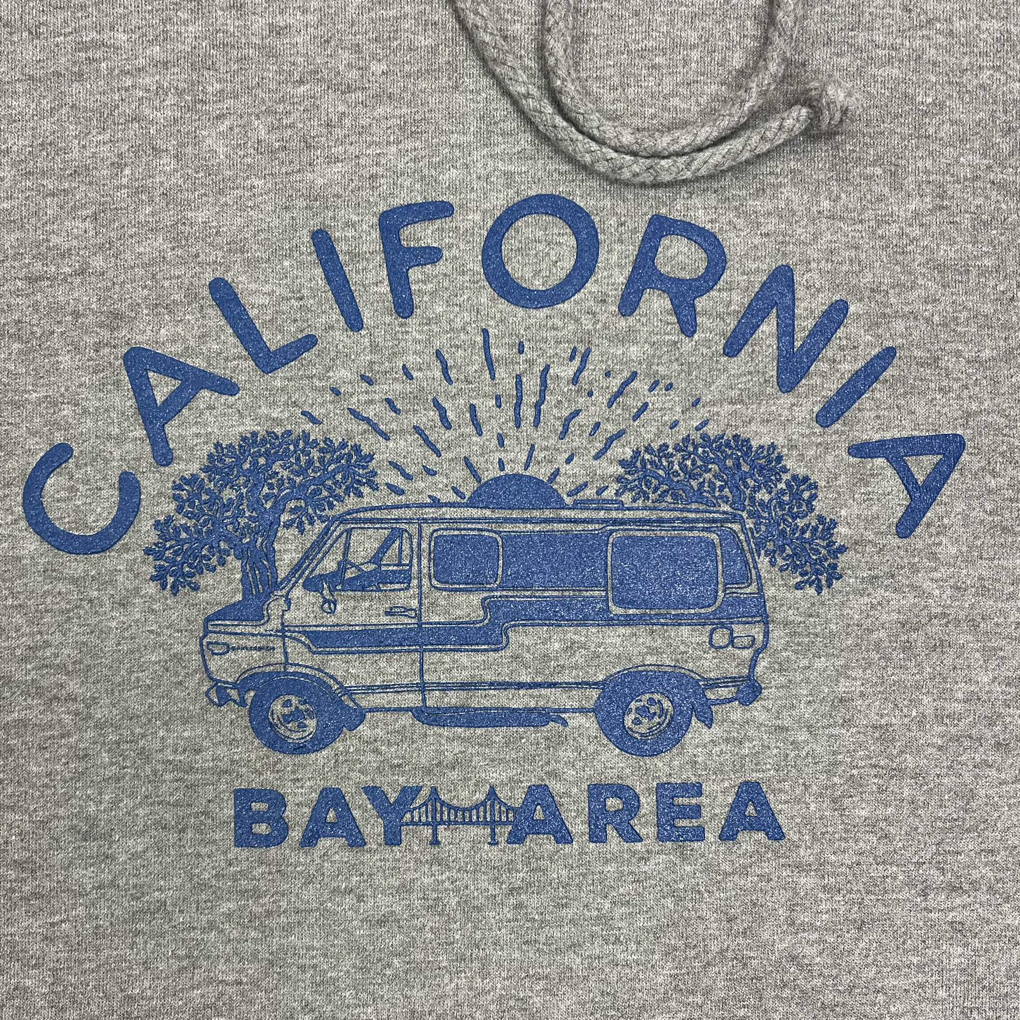 The Wavy Nostalgia Hoodie by Never Elsewhere showcases a gunmetal heather color with a striking blue graphic featuring a van parked under two trees against the backdrop of a setting sun. The design includes the words "CALIFORNIA" above and "BAY AREA" below, incorporating imagery of the iconic Golden Gate Bridge. This pullover hoodie is tailored in men's sizing and includes a practical drawstring hood.