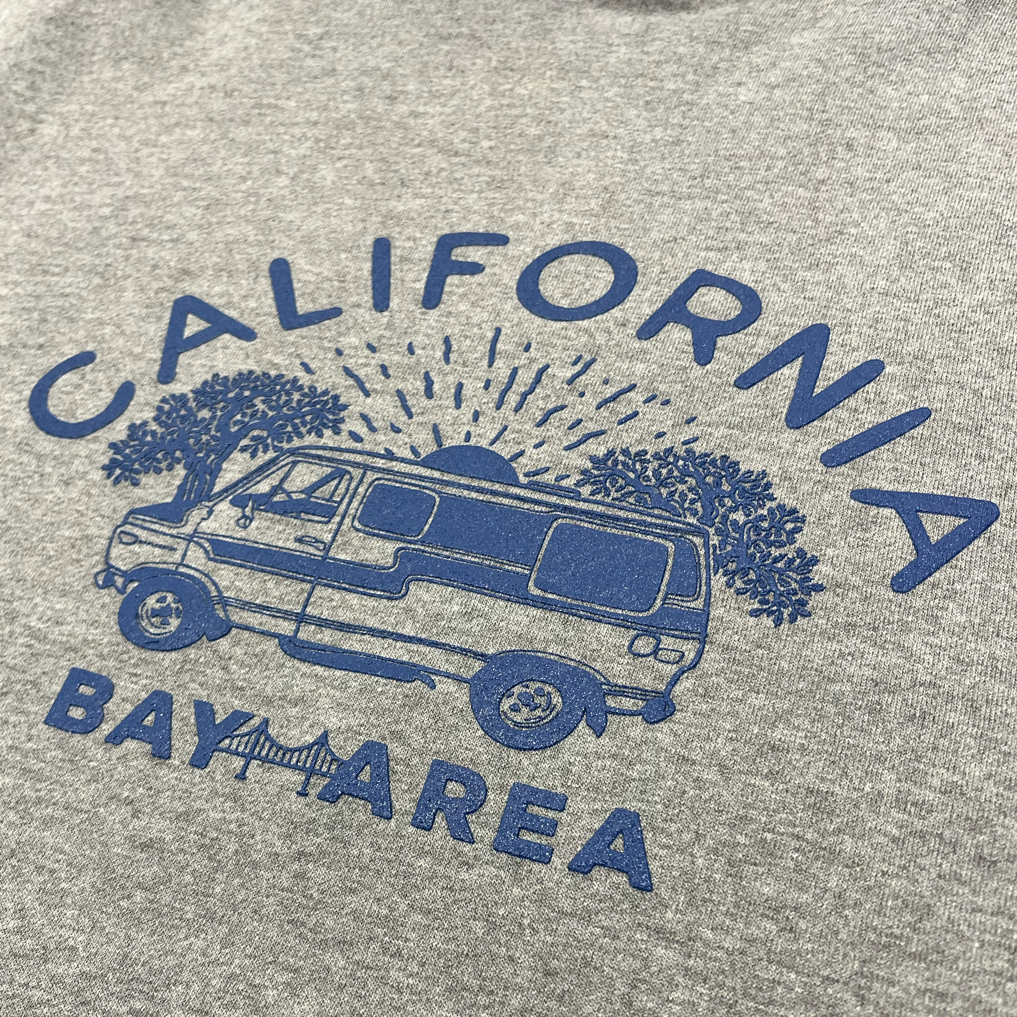 The Wavy Nostalgia Hoodie by Never Elsewhere features a Gunmetal Heather gray pullover design with a blue graphic depicting a van beneath two large trees. A sun with rays shines behind the van, and the words "CALIFORNIA" and "BAY AREA" surround the graphic, with a bridge illustration located below the "BAY AREA" text. Available in men's sizing.