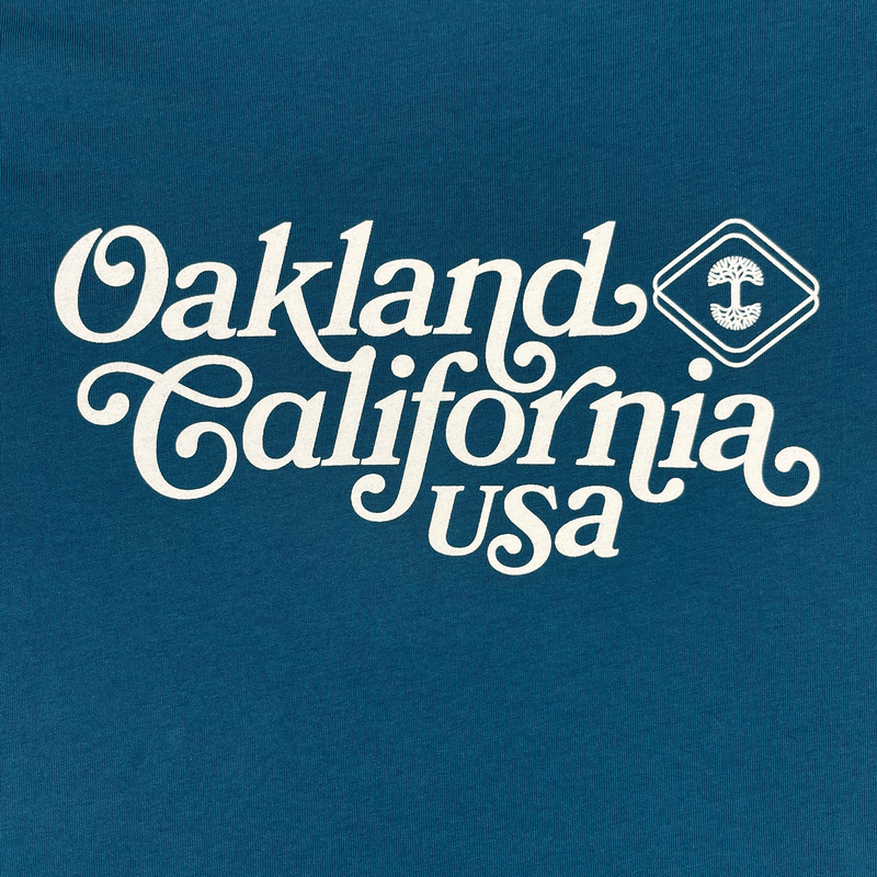 Close-up of Oakland, California, USA graphic in cursive and Oaklandish logo on an atlantic blue t-shirt.