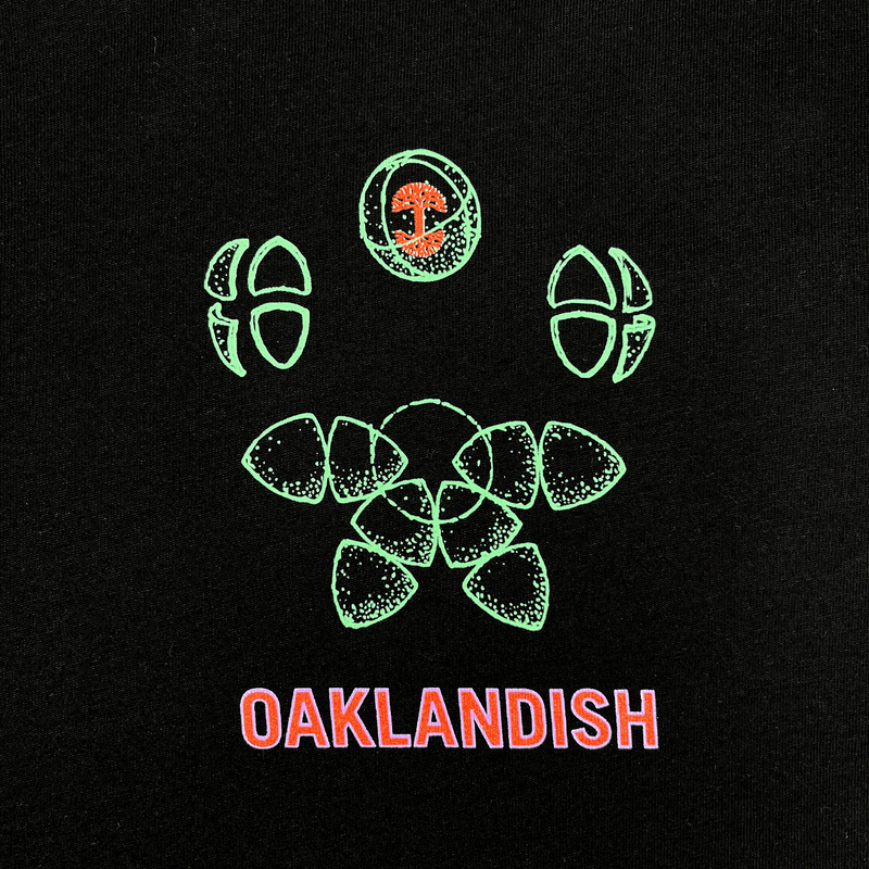 Close-up of green and red Oaklandish worldwide graphic on the left chest wear side of a black t-shirt.