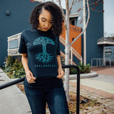 Woman standing outside in a navy t-shirt with a large light blue Oaklandish tree logo and wordmark on the chest.