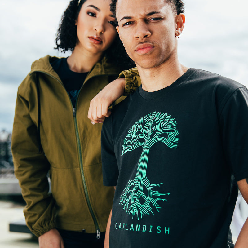 Man and woman standing outside. The woman is in a navy t-shirt with a large light blue Oaklandish tree logo and wordmark on the chest.