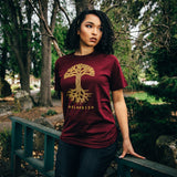 Woman standing outside in a burgundy t-shirt with a large gold Oaklandish tree logo on the chest.