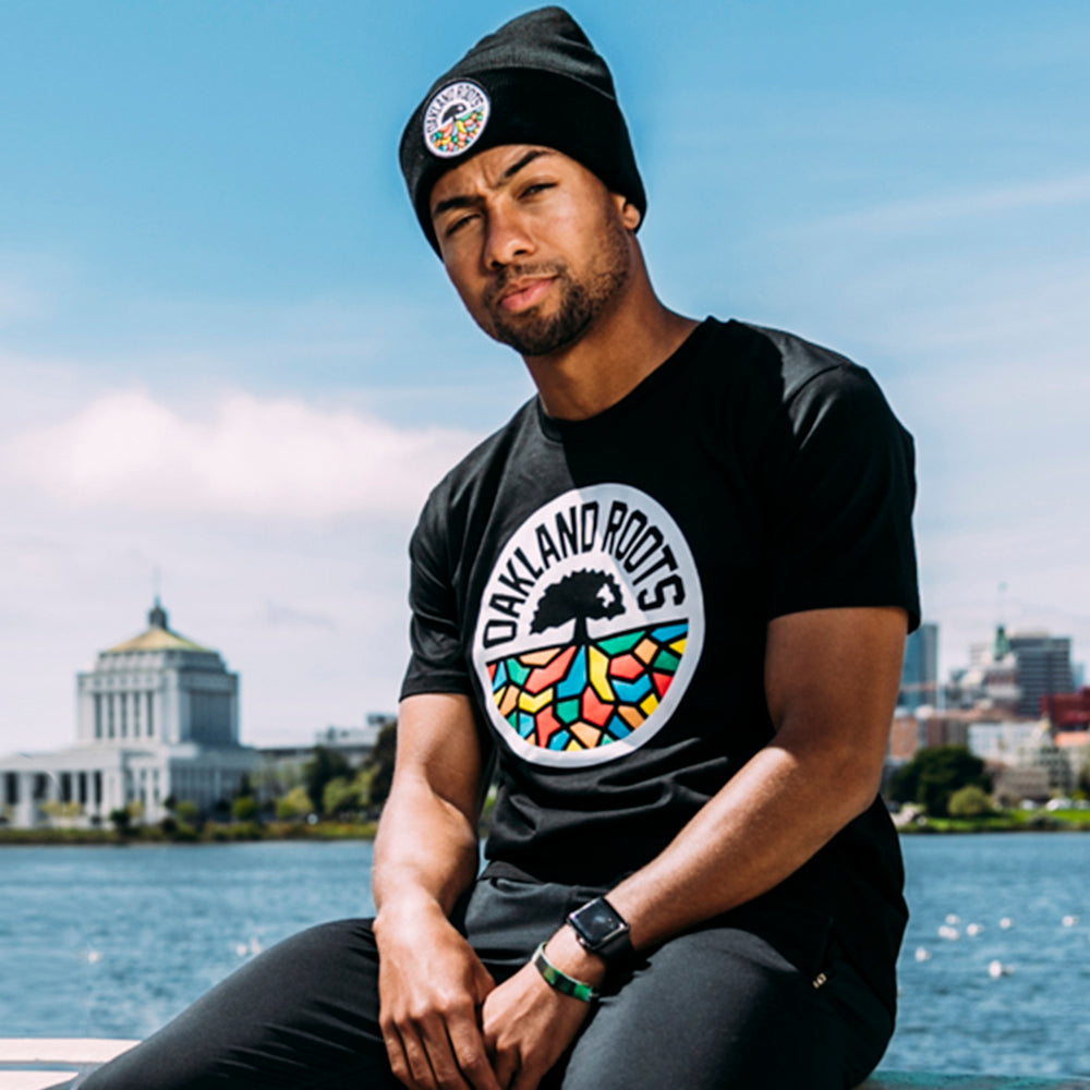 A man sitting outside beside a lake wearing a black beanie and t-shirt with full-color round Oakland Roots SC logos.