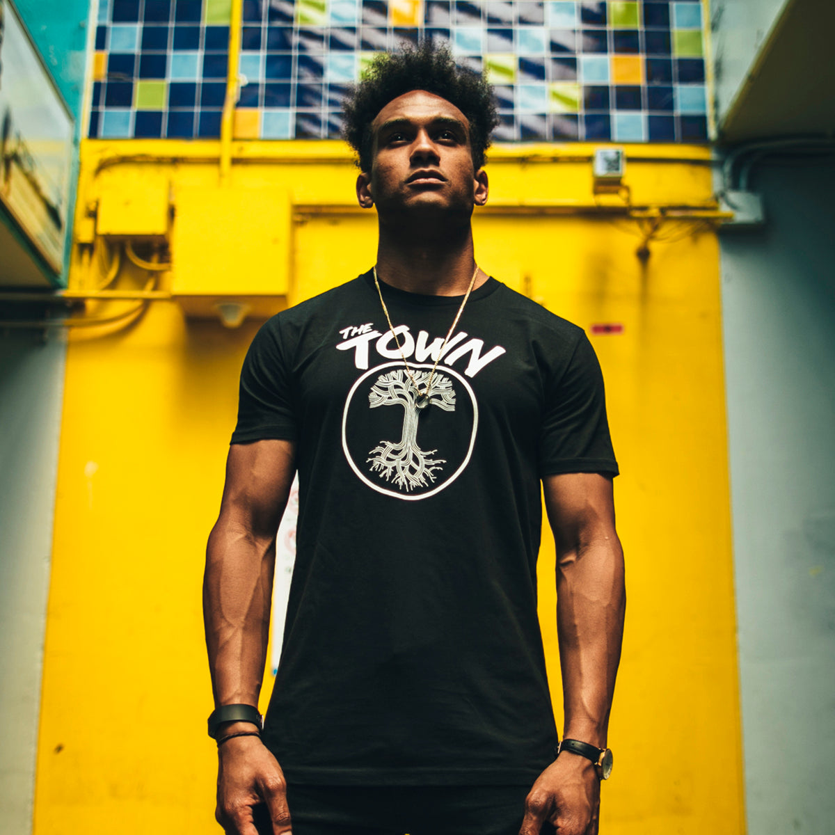 A man stands confidently against a vibrant yellow wall adorned with blue tiles at the top. He wears a black, classic fit Forever Tee from the men's collection by Oaklandish, featuring "The Town" and a tree graphic on the front. His muscular arms are visible, and he accessorizes with a wristwatch and a small necklace. The angle is low, emphasizing his stance.