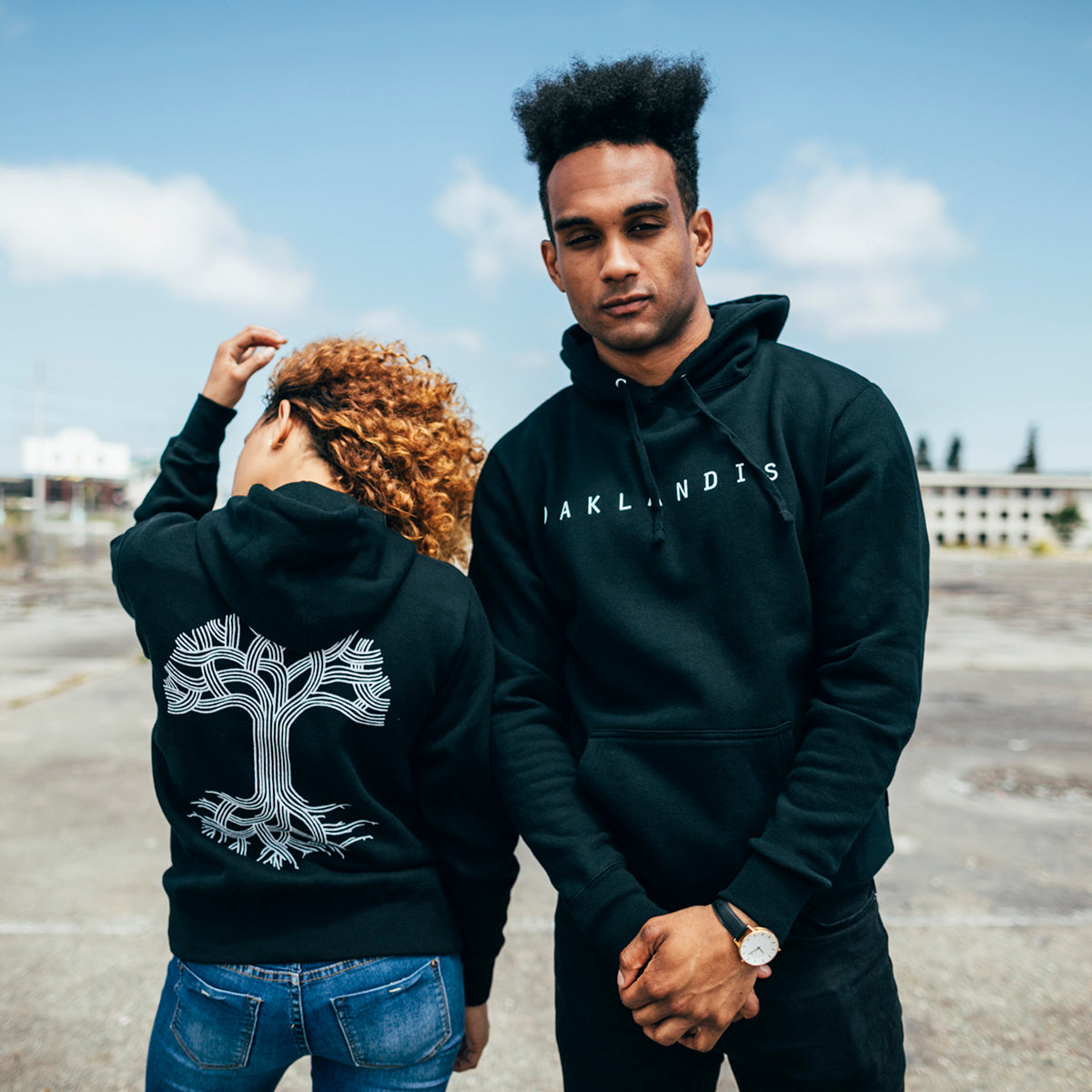 Man & woman outside wearing black hoodies with OAKLANDISH wordmark on the man’s chest and tree logo on the woman’s back.