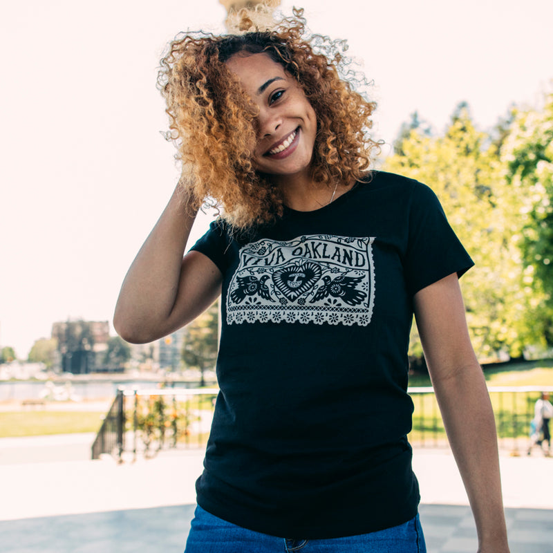 Women standing outdoors in black t-shirt with gold Viva Oakland graphic with hearts and birds on the chest.