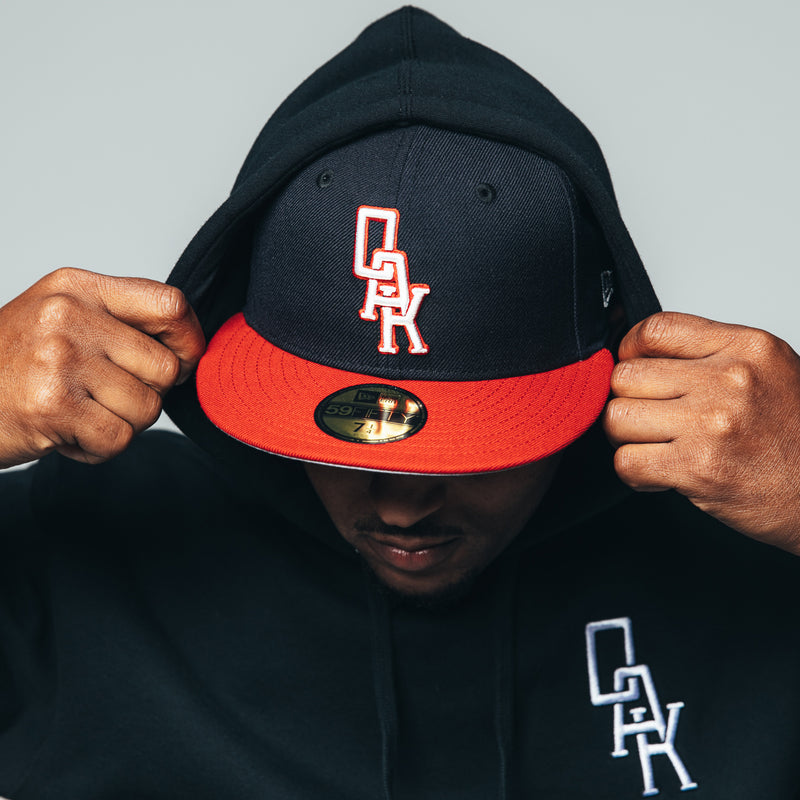 Man wearing a navy New Era cap with red visor and white embroidered OAK wordmark on the crown.