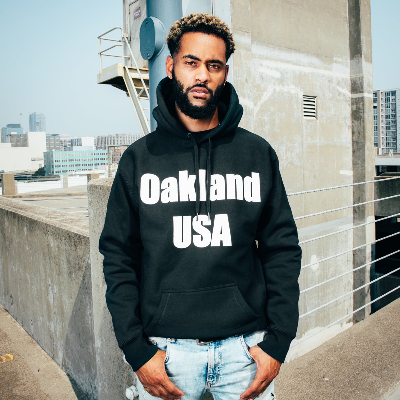 A man standing on an urban rooftop wearing a black hoodie with a white OAKLAND USA wordmark on the chest.