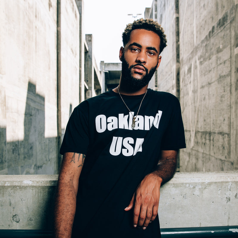 A man in an urban outdoor area wearing a black-t-shirt with a white OAKLAND USA wordmark on the chest.