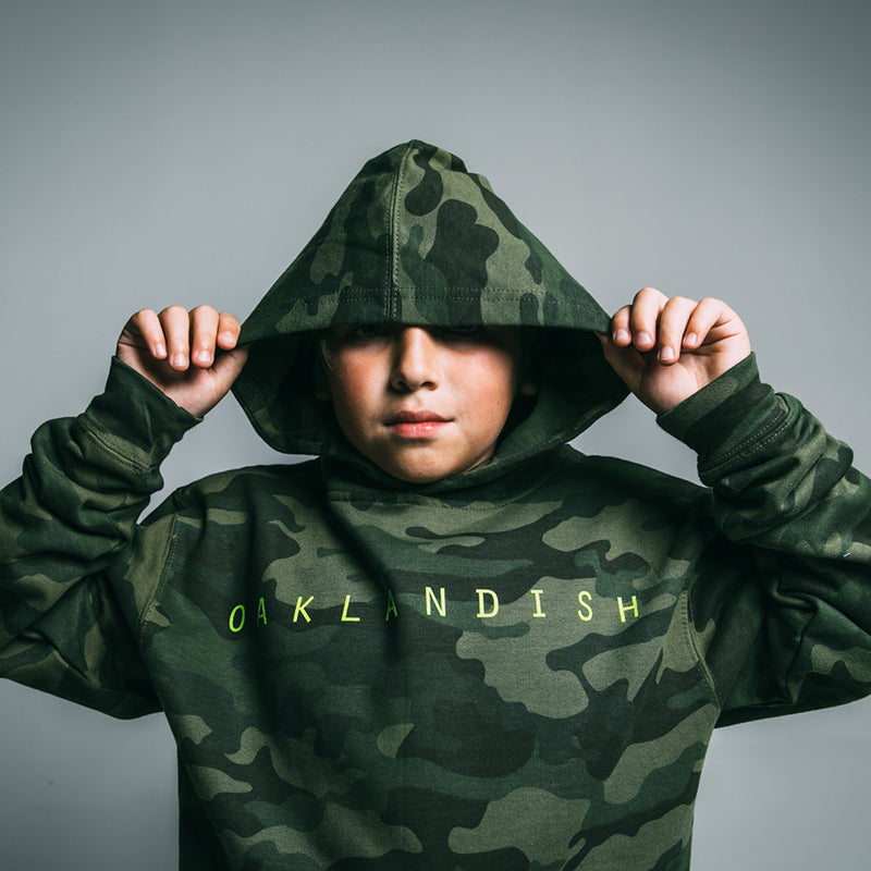 Kid holding the hood of his green camo youth hoodie with light green Oaklandish wordmark on the chest.