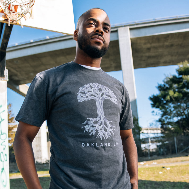 A man standing outside in a charcoal heather t-shirt with a large white Oaklandish tree logo on the chest.