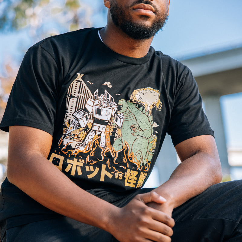 A man’s chest outdoors with a graphic depiction of Kaiju vs. Autobart monsters invading Oakland on a black t-shirt. 