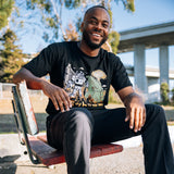 A smiling man sitting outdoors wearing a black t-shirt with a graphic of Kaiju vs. Autobart monsters invading Oakland. 