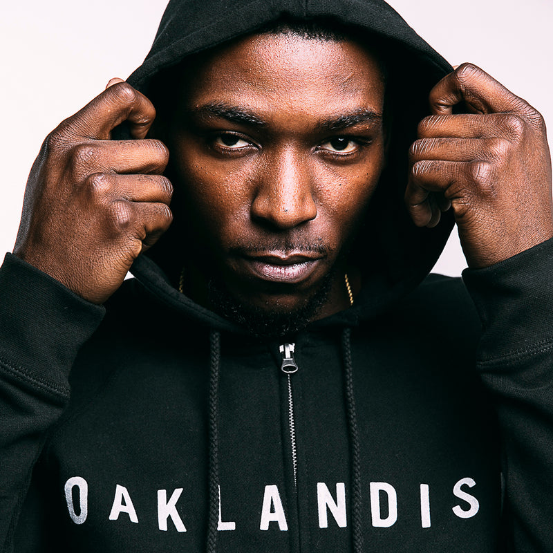 Close up of man’s face with hands holding the hood of a black zip-up hooded sweatshirt with white Oaklandish wordmark on chest.