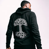 A man standing back to the camera displaying the backside of a black hooded sweatshirt with a large white Oaklandish tree logo.