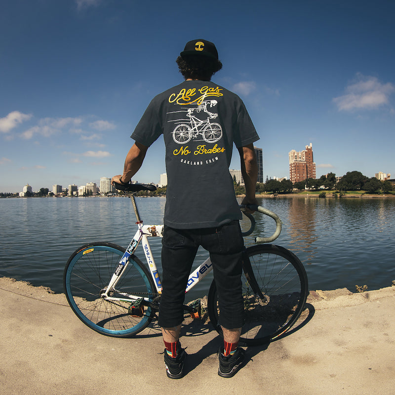 A man standing in front of a lake with a bike wearing a black t-shirt with All Gas No Breaks, Oakland Crew design.
