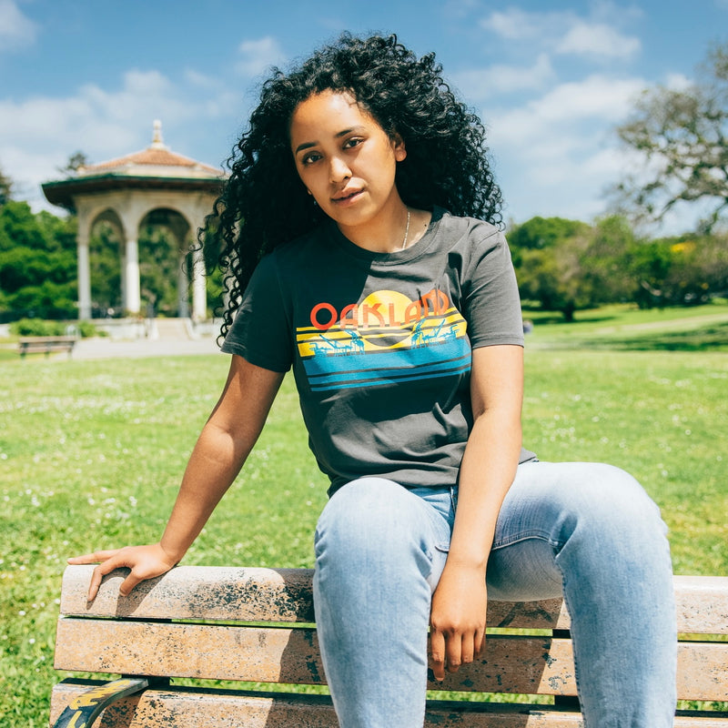 Woman sitting outside in a faded black women’s t-shirt with multi-color license plate design with capital letters spelling out OAKLAND.