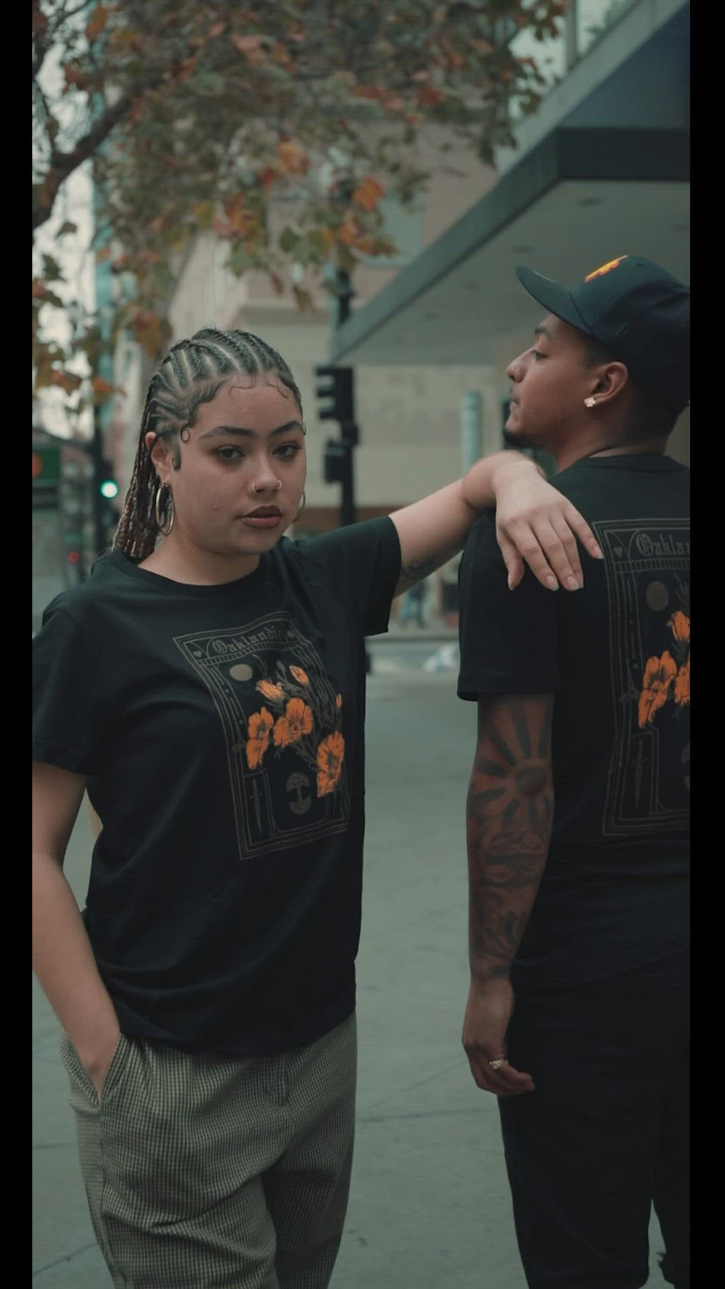 A man and woman standing on an Oakland street wearing black Oaklandish Blossom t-shirts.