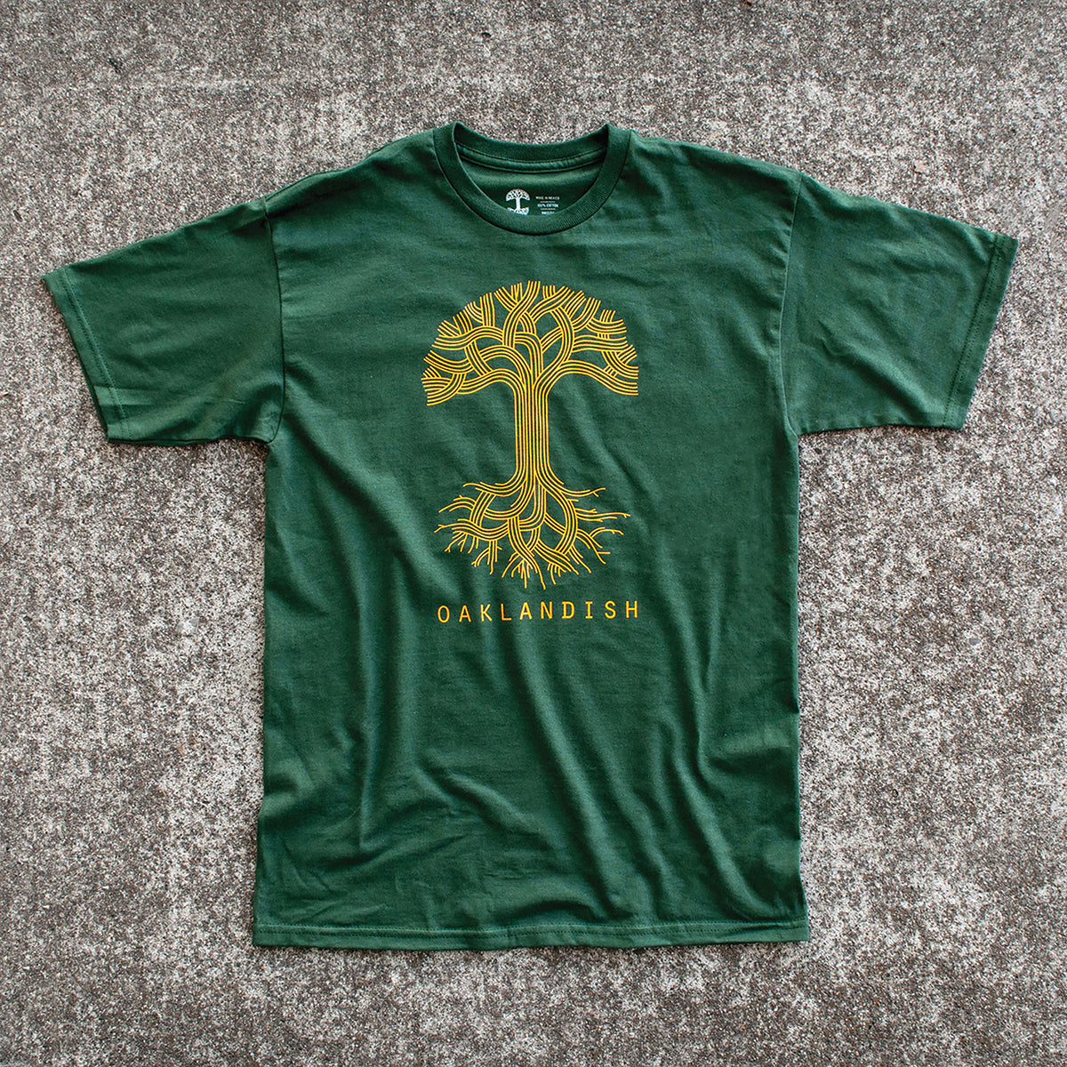 An Oaklandish Classic Logo Tee in dark green is laid flat on a gray surface. Made from 100% cotton, this shirt showcases a golden-yellow graphic of a stylized tree with intricate branching patterns above and below the ground. Below the tree graphic, matching golden-yellow text reads "OAKLANDISH.