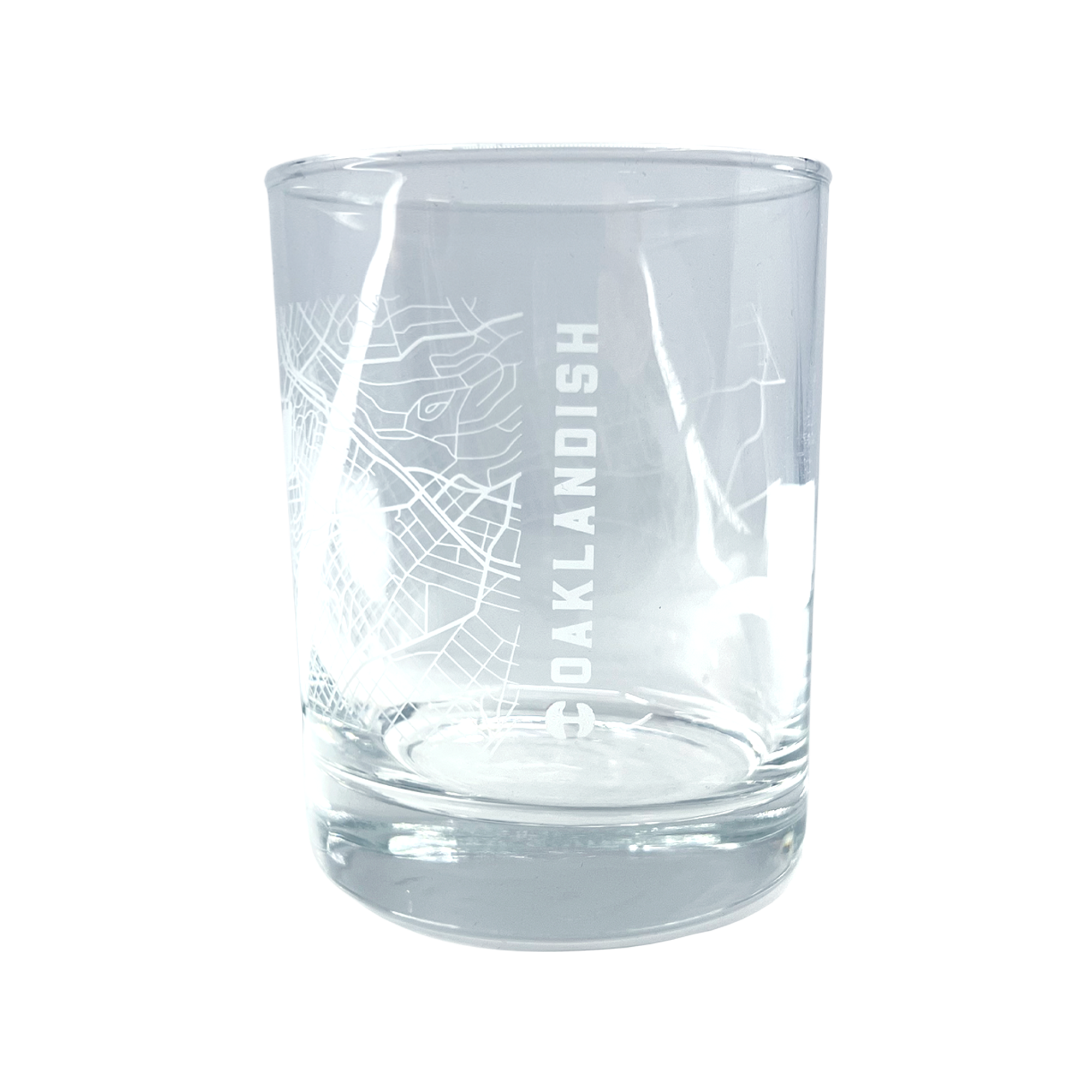 The Aerial Tumbler by Oaklandish is a clear glass whiskey tumbler with a minimalist design that boasts an etched, abstract aerial map of Oakland. The word "OAKLANDISH" is prominently displayed in vertical capital letters down one side of the classic cylindrical glass, which features a thick base.