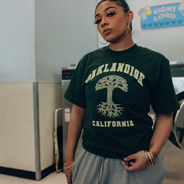 Female model wearing ivy green t-shirt with creamy yellow Oaklandish California wordmark and Classic Oaklandish tree logo centered while washing in laundry mat.