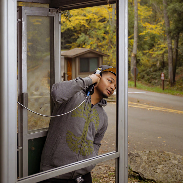 A man leaning out of a phone booth on a wooded street wearing a grey zip-up hoodie with olive Oaklandish tree appliqué on the chest.