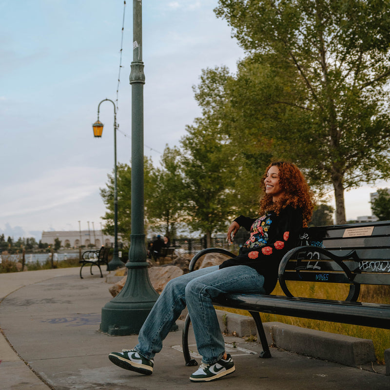 Women sitting on an urban park bench in a long sleeve black t-shirt featuring flowers and birds designed by artist Jet Martinez. 