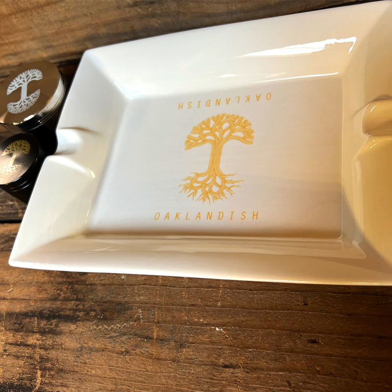 White ceramic ashtray with yellow Oaklandish tree logo and wordmark in the center on wood table with two grinders.