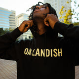 Man outside in Oakland wearing black pullover hoodie with yellow Oaklandish wordmark on the chest and tree logo on sleeve.