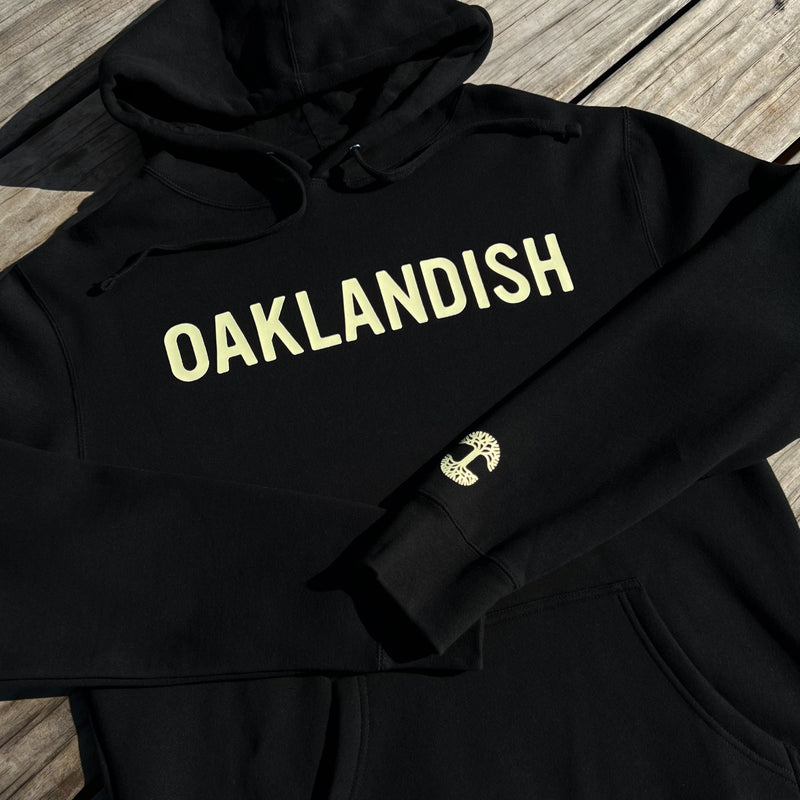 Black hoodie with yellow Oaklandish wordmark on chest and arm folded over to show tree logo on sleeve outdoors on wood deck.