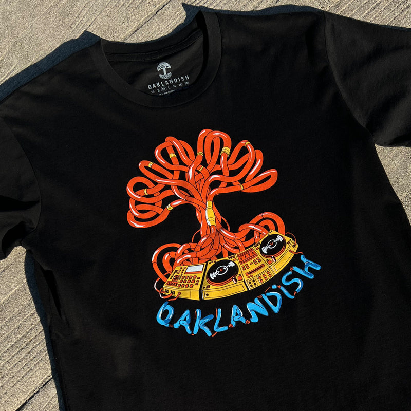 Black t-shirt with Oaklandish DJ crew graphic with a turntable, wires & Oaklandish wordmark in orange, yellow and blue on wood deck.