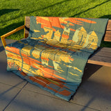 Puffy blanket with Oakland cityscape in blue, gold, rust and white laying over outdoor bench at sunset.