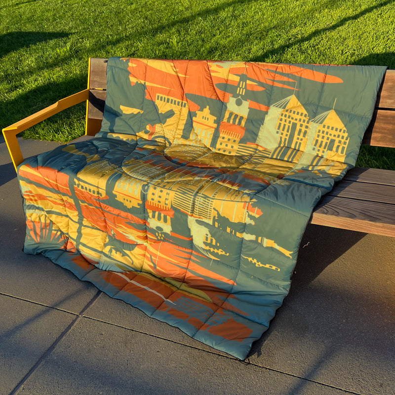 Puffy blanket with Oakland cityscape in blue, gold, rust and white laying over outdoor bench at sunset.