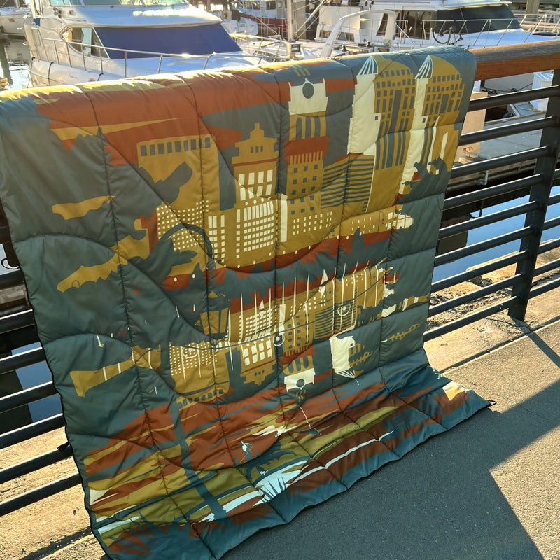 Puffy blanket with Oakland cityscape in blue, gold, rust and white laying over a marina fence at sunset.