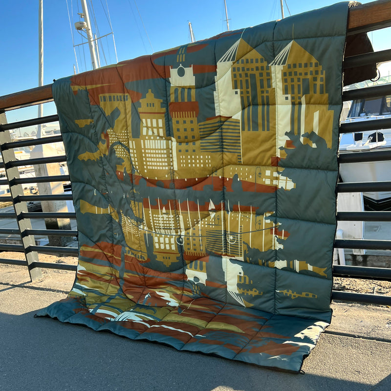Puffy blanket with Oakland cityscape in blue, gold, rust and white laying over a marina fence at sunset.