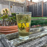 Clear glass pint beer glass with gold OAK wordmark in a bleed font, full of beer sitting on wood shelf in a backyard.