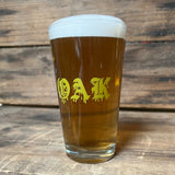 Clear glass pint beer glass with gold OAK wordmark in a bleed font, full of beer sitting on wood shelf.