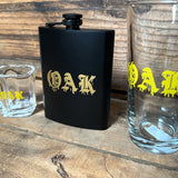 Black flask with gold OAK wordmark in a bleeding font on the wood shelf with glass shot glass and pint glass with the same wordmark.