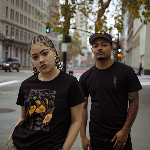A man and woman standing on an Oakland street wearing black Oaklandish Blossom t-shirts.