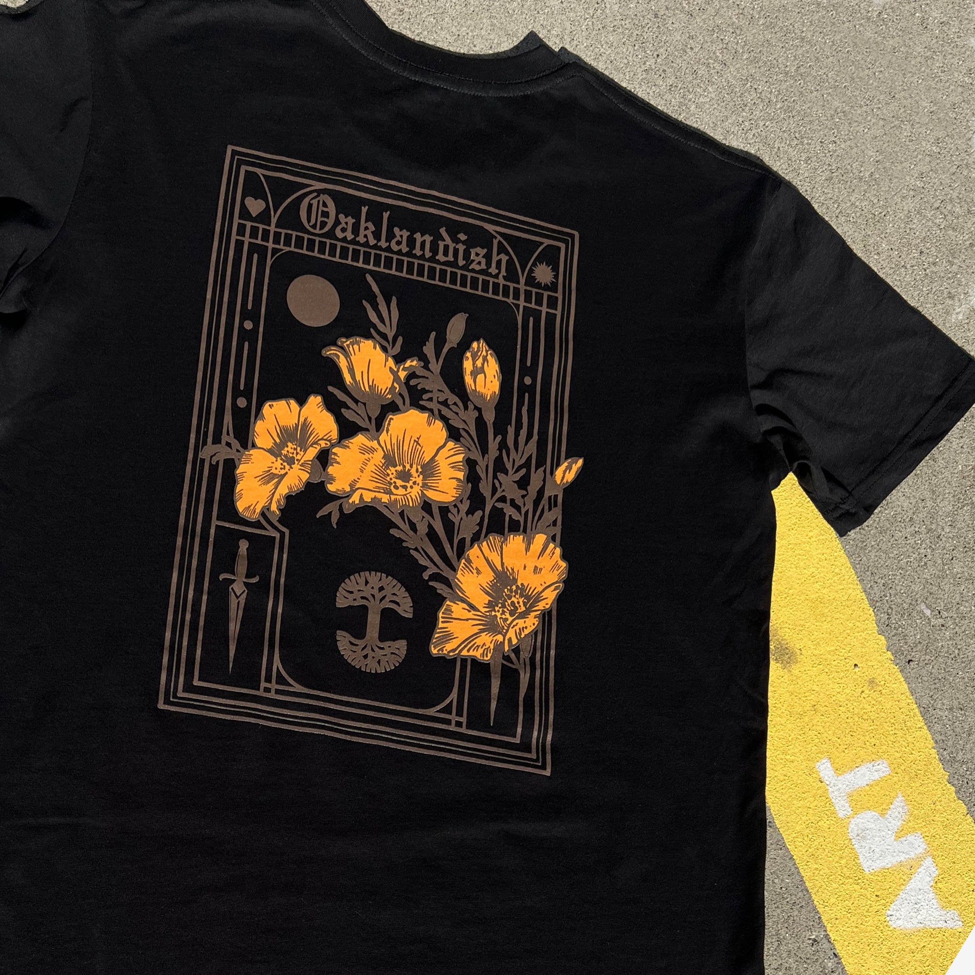 The back of a black cotton t-shirt with California poppies, an Oaklandish wordmark, and tree logo lying on the street.