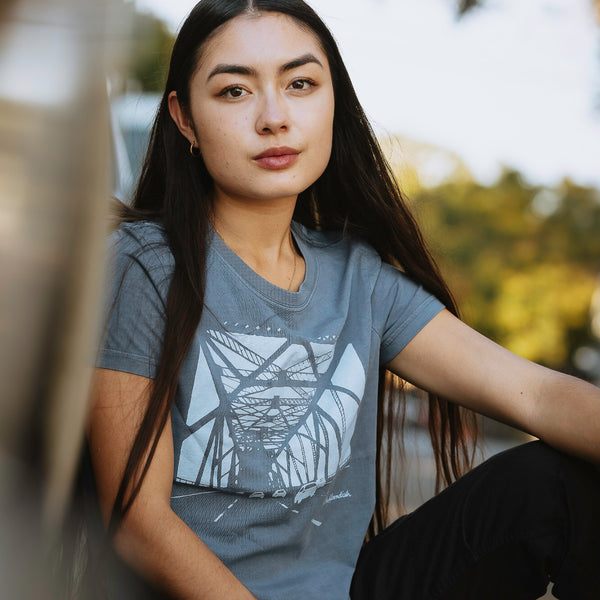 Female model outside wearing Faded blue women’s t-shirt with a graphic of the eastern span of Oakland bridge.