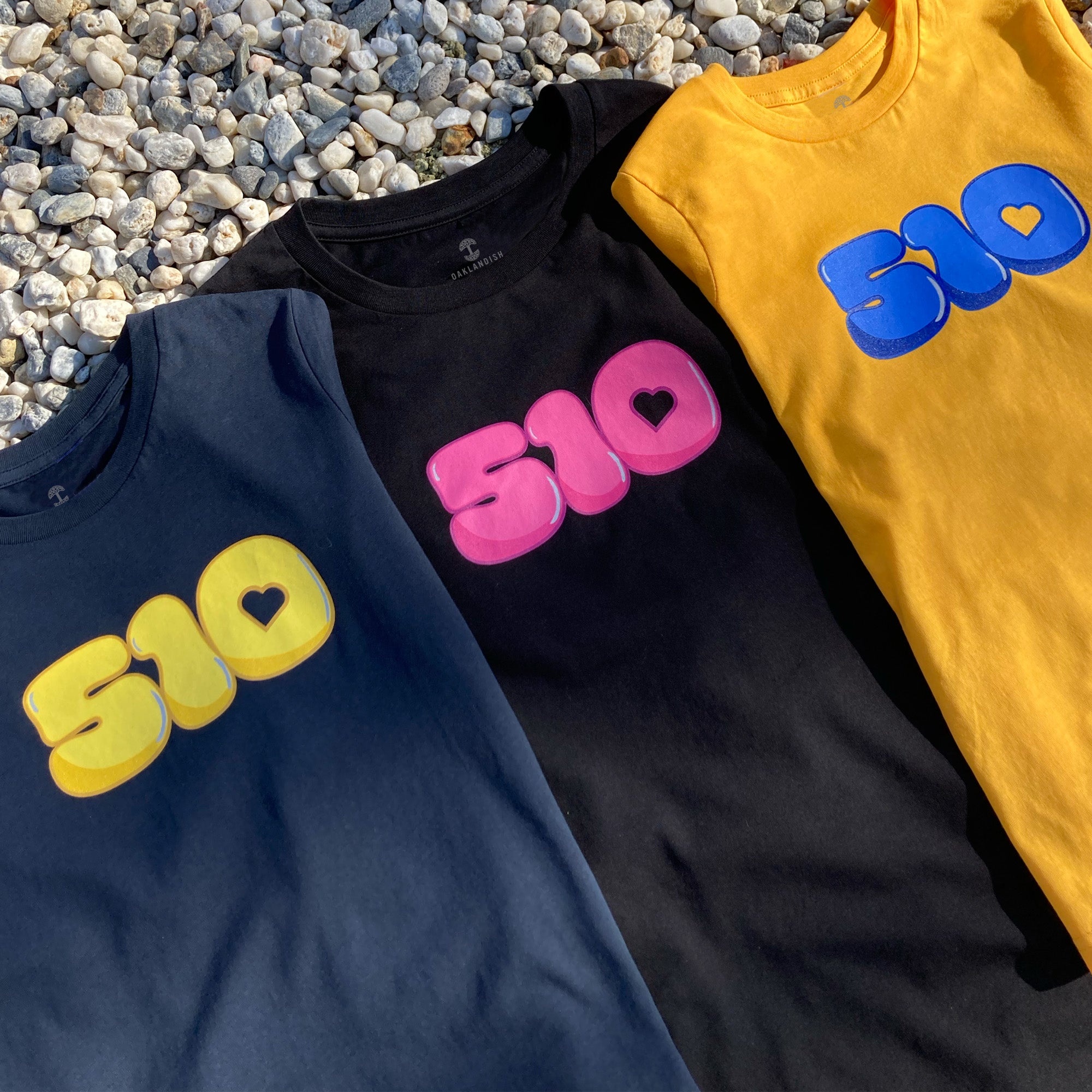 Three Youth Bubble 510 Tees from Oaklandish are laid out on a bed of white pebbles, each adorned with a "510" design featuring a heart within the "0." The first T-shirt is navy blue with yellow text, the second is black with pink text, and the third is mustard yellow with blue text. Made from 100% cotton, all shirts have a casual style with short sleeves.