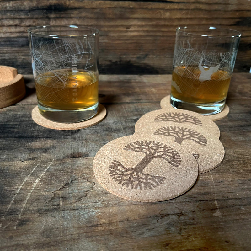Set of 6 round cork coasters with a darker brown Oaklandish tree logo spread out on a wood table, two with a glass of whiskey. 